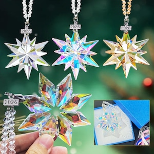 May include: A collection of star-shaped ornaments with iridescent crystal facets. Each ornament is suspended by a chain and features the year "2026". One ornament is held by a hand, and another is displayed in a blue box.