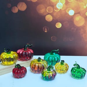 May include: A selection of colourful glass pumpkins in shades of yellow, red, green, and orange. Each pumpkin has a dark green stem. The pumpkins are arranged on a white surface with a blurred background.