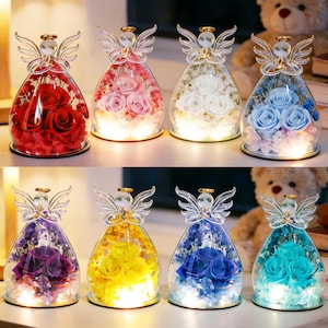 Pode incluir: Uma coleção de figuras de anjos de vidro, cada uma preenchida com flores preservadas coloridas. Os anjos têm asas e halos de cristal. As flores incluem rosas vermelhas, rosas, brancas, azuis, roxas, amarelas e turquesas, com luz quente.