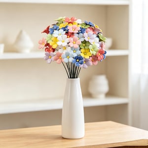 Op de afbeelding: Een boeket van veelkleurige keramische bloemen met gele harten, elk bevestigd aan een dunne zwarte draadsteel. De bloemen zijn gerangschikt in een hoge, gladde, witte keramische vaas met een taps toelopende hals en een brede, platte voet.