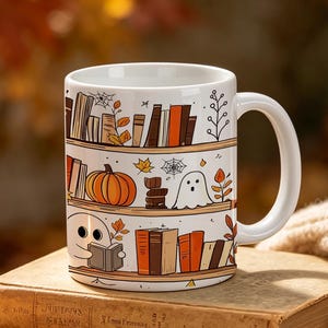 May include: A white ceramic mug featuring a wrap-around illustration of wooden shelves filled with books, a pumpkin, and small ghosts. The design includes autumn leaves and spider webs in shades of orange, brown, and black on a white background.