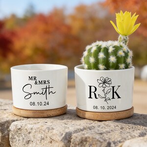 Puede incluir: Dos macetas de cerámica blanca con bases de bambú. La maceta izquierda presenta el texto MR & MRS Smith y 08.10.24. La maceta derecha contiene un cactus verde con una flor amarilla y presenta el texto R K con un icono floral y 08.10.24.