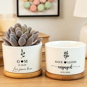 Può includere: Due vasi cilindrici in ceramica bianca con sottovasi in bambù. Uno contiene una pianta succulenta e riporta il testo G M, 11.18.25, e Love grows here. L'altro riporta il testo ALICE LLOYD, engaged, e 6/21/2025.
