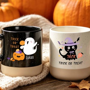 Könnte beinhalten: Zwei Keramikbecher mit zweifarbigem Design. Der schwarze Becher zeigt ein weißes Gespenst, einen orangefarbenen Kürbis sowie den Text "TRICK OR TREAT" und "Lukas". Der weiße Becher zeigt eine schwarze Katze mit lila Hut und den Text "JACK" und "TRICK OR TREAT".