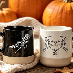 May include: Two ceramic mugs with a two-tone design. The black mug features a white skeleton hand holding a rose and the letters S+K. The cream mug displays two skeleton hands forming a heart shape with the text 'Till Death Do Us Part' and J+L.
