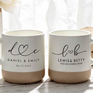 Puede incluir: Dos tazas de cerámica blanca con textura moteada y base color canela. La primera taza tiene las letras d y e con un corazón y el texto DANIEL & EMILY 06.12.2025. La segunda tiene l y b con un corazón y LEWIS & BETTY 9TH DECEMBER.2025.