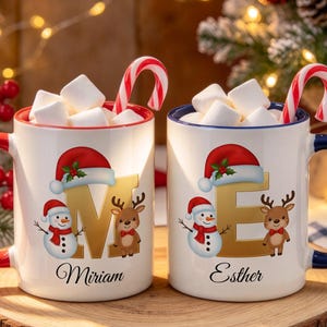 May include: Two white ceramic mugs filled with marshmallows and candy canes. The left mug has a red handle, the letter M, a snowman, a reindeer, and the name Miriam. The right mug has a blue handle, the letter E, a snowman, a reindeer, and the name Esther.