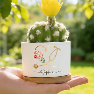 May include: A white ceramic pot with a cactus and a yellow flower. The pot features a gold stethoscope and floral design with the name "Sophia" in script. The pot sits on a wooden saucer.