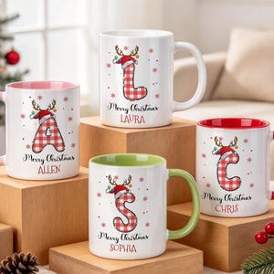 Könnte beinhalten: Vier weiße Keramiktassen mit farbigem Innenraum und Henkel. Jede Tasse zeigt einen rot-weiß karierten Buchstaben mit einem Rentiergeweih-Weihnachtsmannmütze, roten Schneeflocken sowie dem Text Merry Christmas und einem personalisierten Namen.