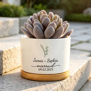 Puede incluir: Una maceta cilíndrica de cerámica blanca con base de platillo de bambú, que contiene una suculenta de color gris verdoso. La maceta presenta un gráfico de rama verde sobre el texto negro: James + Sophia married 09.02.2025.
