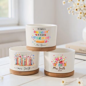 Puede incluir: Tres macetas de cerámica blanca con bases de madera. Una maceta tiene el texto "IT TAKES A BIG HEART TO TEACH LITTLE MINDS" y un gráfico de lápiz. Otra presenta un diseño floral con libros. La tercera maceta dice "TEACHER Mrs. Smith 2025".