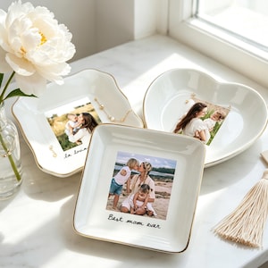 May include: Three white ceramic trinket dishes with gold rims, each holding a framed photograph. One dish is square, another heart-shaped, and the third has an irregular shape. The square dish has the text "Best mom ever". A white tassel hangs nearby.