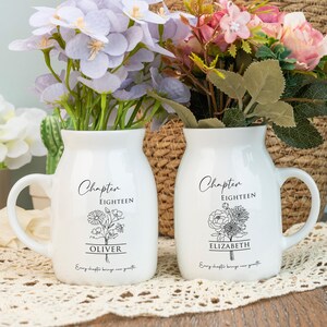 May include: Two white ceramic vases with handles, each holding colorful flowers. The vases have the text "Chapter EIGHTEEN" and a name, "OLIVER" and "ELIZABETH", with floral illustrations. The phrase "Every chapter brings new growth" is also printed on the vases.