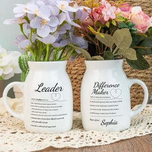 Puede incluir: Dos jarrones de cerámica blanca con asas, cada uno con flores. Uno dice "Leader" con una definición y el nombre "Sophia". El otro dice "Difference Maker" con una definición y el nombre "Sophia".