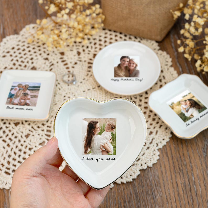 May include: A collection of white ceramic trinket dishes with gold trim, each featuring a photograph. One dish is heart-shaped with "I love you nana" text. Other dishes include "Best mom ever" and "Happy Mother's Day!" text.