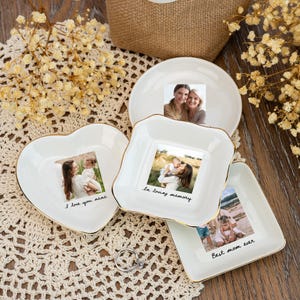 May include: A collection of white ceramic dishes in various shapes, including a heart, square, and round, with gold trim. Each dish features a photograph and a handwritten message. The dishes are arranged on a lace doily with dried flowers.
