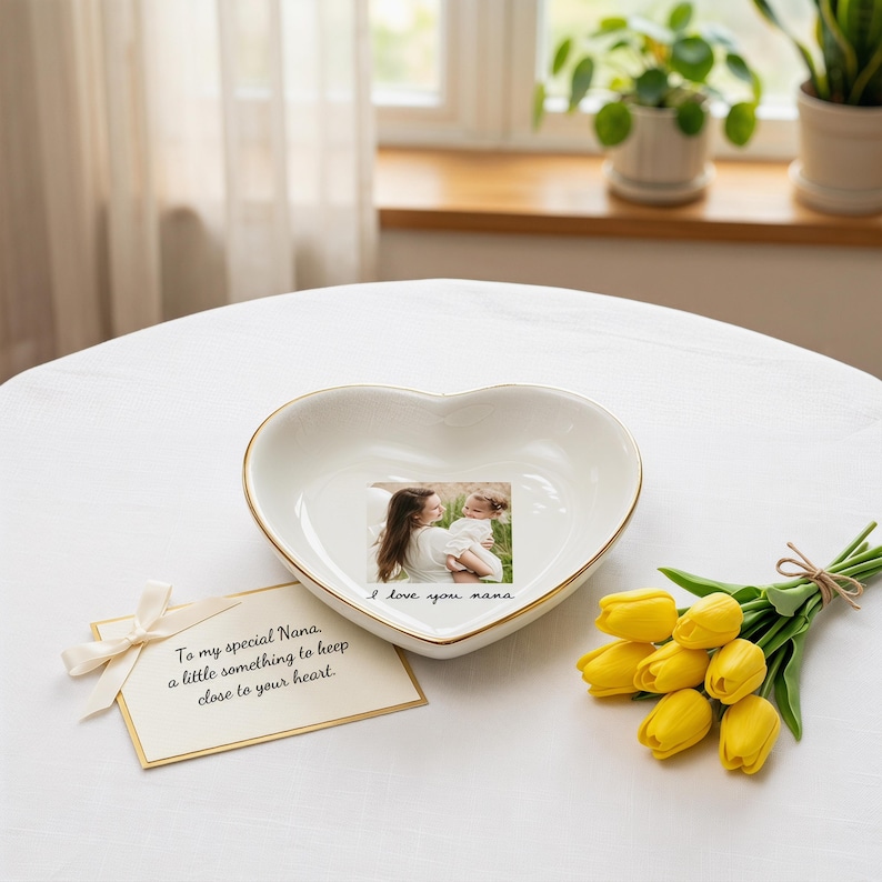 May include: A heart-shaped, white ceramic dish with a gold rim, featuring a photo and the words "I love you nana." A card with a bow reads "To my special Nana, a little something to keep close to your heart." Yellow tulips are arranged nearby.