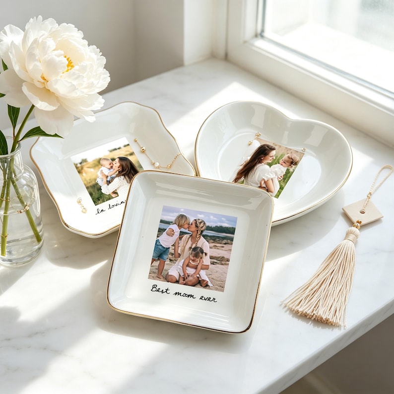 May include: Three white ceramic trinket dishes with gold rims, each holding a framed photograph. One dish is square, another heart-shaped, and the third has an irregular shape. The square dish has the text "Best mom ever". A white tassel hangs nearby.