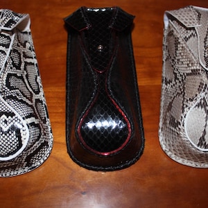 May include: Three eyeglass cases in a row, each with a unique design. The case on the left has a black and white snakeskin pattern, the middle case is black with red trim, and the case on the right has a brown and tan snakeskin pattern.