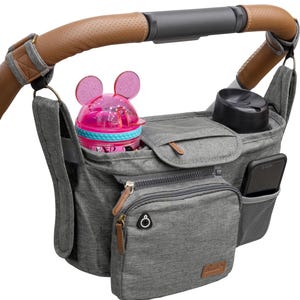 May include: Gray stroller organizer with multiple pockets, including a zippered pocket, a mesh pocket, and a large open pocket. A pink sippy cup with Mickey Mouse ears is in the open pocket, and a black travel mug is in the mesh pocket.
