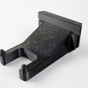 Könnte beinhalten: Ein schwarzer, 3D-gedruckter Wandhalter mit strukturierter Oberfläche. Die Halterung hat eine schräge Ablage und zwei gebogene Arme mit abgerundeten Spitzen. Das Design ist modern und minimalistisch.