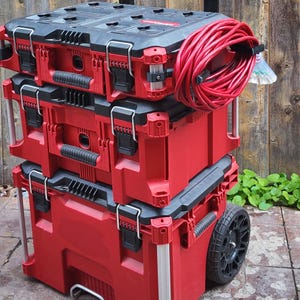 May include: A stack of red and black toolboxes with a red extension cable. The toolboxes are on a rolling cart with black wheels and a silver handle. The toolboxes have black latches and handles.