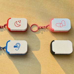 May include: Four rectangular keyrings with rounded edges in red, pink, blue, and black. Each keyring has a white face with a different icon: a crescent moon with Zzz's, two pills, an ear, and a blank one. The keyrings are on a light brown surface.