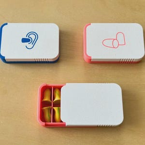 Puede incluir: Tres estuches rectangulares para tapones para los oídos. Uno es blanco con un borde azul y un gráfico de oreja. Otro es blanco con un borde rosa y gráficos de tapones para los oídos. El tercero está abierto, mostrando tapones para los oídos amarillos dentro de un estuche rosa.