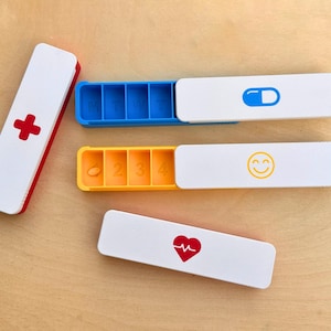Op de afbeelding: Witte en rode pillenorganizers met blauwe, gele en witte compartimenten. Eén heeft een rood kruis, een ander een blauw pil-icoon, een gele smiley en een rood hartsymbool.