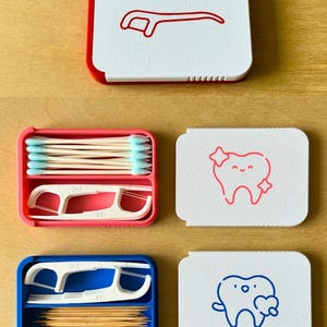 May include: A selection of dental hygiene products, including floss picks, cotton buds, and toothpicks, in colourful, compact cases. The cases are white with red, pink, and blue accents. Each case has a unique design, including a floss pick and a smiling tooth.