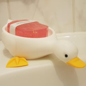 Könnte beinhalten: Ein weißer, entenförmiger Seifenschwamm mit einem rosa und weißen Stück Seife darin. Der Schwamm hat gelbe Füße und einen Schnabel.