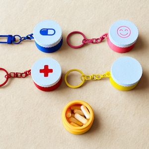 Pode incluir: Uma coleção de chaveiros coloridos para recipientes de comprimidos. Cada chaveiro apresenta um recipiente redondo com uma tampa branca e uma base colorida. As tampas têm símbolos diferentes: uma pílula azul, uma cruz vermelha e uma carinha sorridente. Um recipiente está aberto, cheio de comprimidos.
