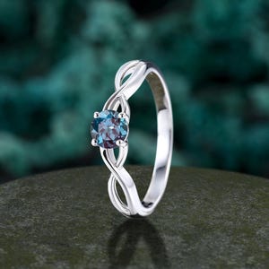 May include: A silver ring with an infinity-style band, featuring a round, dark blue gemstone. The ring is set against a blurred, teal-green background, highlighting the polished metal and the gem's color.
