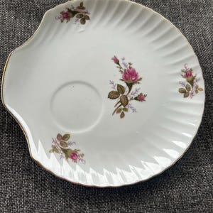 May include: A white porcelain saucer with a scalloped, ribbed edge and a gold-trimmed rim. The surface features a central circular indentation and is decorated with small, delicate floral motifs of pink rosebuds and green leaves.