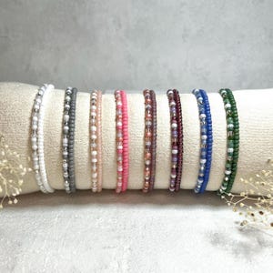Könnte beinhalten: Eine Sammlung von Armbändern mit Perlen in verschiedenen Farben, darunter Weiß, Grau, Rosa, Lila, Blau und Grün. Die Armbänder werden auf einem cremefarbenen Kissen präsentiert, das ihr zartes Design und ihre lebendigen Farbtöne hervorhebt. Jedes Armband besteht aus kleinen, farbigen Perlen.