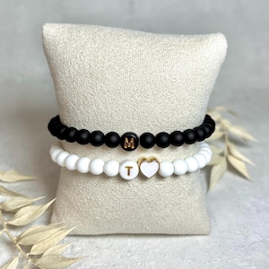 Könnte beinhalten: Zwei Perlenarmbänder, eines in Schwarz und eines in Weiß, werden auf einem cremefarbenen Kissen präsentiert. Das schwarze Armband hat eine Perle mit dem Buchstaben "M". Das weiße Armband hat einen herzförmigen Anhänger und eine Perle mit dem Buchstaben "T".