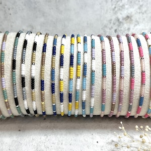 Könnte beinhalten: Eine Sammlung von 18 bunten Perlenarmbändern, jedes mit einem anderen Muster aus blauen, weißen, gelben, rosa und grauen Perlen. Die Armbänder sind in einer Reihe auf einer weißen Oberfläche angeordnet.
