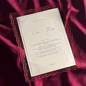 Può includere: Un invito di nozze con un bordo in pizzo bordeaux e un biglietto bianco con calligrafia. L'invito è ambientato su uno sfondo di raso bordeaux. Il testo include nomi e una data.
