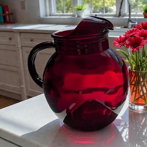 May include: A large, ruby-red glass pitcher with a rounded body and a handle. Beside it, a small glass vase holds a bouquet of bright pink flowers. The pitcher sits on a white countertop in a well-lit kitchen setting.