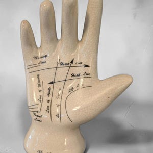 May include: A cream-colored ceramic hand sculpture with a crackle glaze finish. The palm features black printed text and lines labeled Marriage Lines, Heart Line, Head Line, Life Line, Line of Mercury, Line of Apollo, Line of Saturn, and Travel Lines.