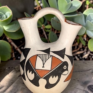 May include: A cream-colored ceramic vessel featuring two spouts connected by a central arched handle. The exterior is decorated with geometric patterns in black and terracotta, including stylized shapes and fine vertical lines on the rounded body.