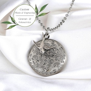 May include: A silver-toned locket necklace with a bird charm. The round locket features an engraved floral design and a small bird. The necklace includes the text "Custom Photo & Engraving" and "Gravur- & Fotoservice".
