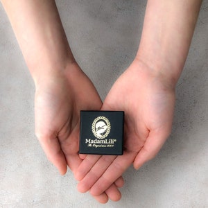 May include: A small, black box with gold lettering and a decorative emblem is held in open hands. The box reads "MadamLili" and "The Original since 2009". The background is a light gray, textured surface.