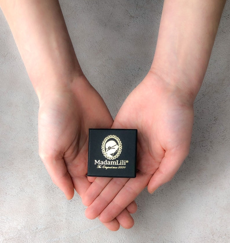 May include: A small, black box with gold lettering and a decorative emblem is held in open hands. The box reads "MadamLili" and "The Original since 2009". The background is a light gray, textured surface.