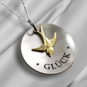 May include: A silver necklace with a round pendant featuring a gold bird and the word "GLÜCK" engraved on it.