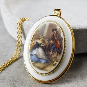 May include: A gold necklace with a white oval pendant featuring a painted scene of a woman in a blue dress and a man in a red and gold outfit. The pendant has a gold rim and a white background.