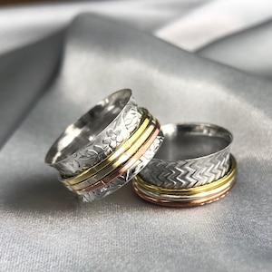 Könnte beinhalten: Zwei silberne Spinnerringe mit dekorativen Mustern. Ein Ring hat ein Blumenmuster und der andere ein Zickzackmuster. Beide Ringe haben drei Bänder aus Gold und Kupfer.