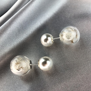 Può includere: Un paio di orecchini a doppia faccia. Un lato presenta una grande perla bianca, e l'altro lato è un globo di vetro trasparente contenente delicato materiale vegetale essiccato. Gli orecchini sono posti su uno sfondo grigio serico.