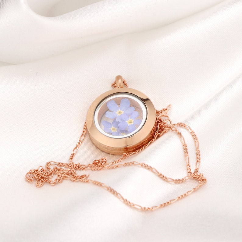 Puede incluir: Un collar con un medall&oacute;n de oro rosa que contiene flores de nomeolvides prensadas en un inserto de vidrio transparente.