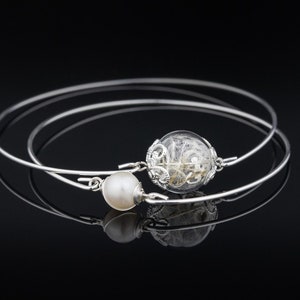 May include: Two silver bangle bracelets with a pearl and a glass orb containing dried flowers. The bracelets are stacked on top of each other.